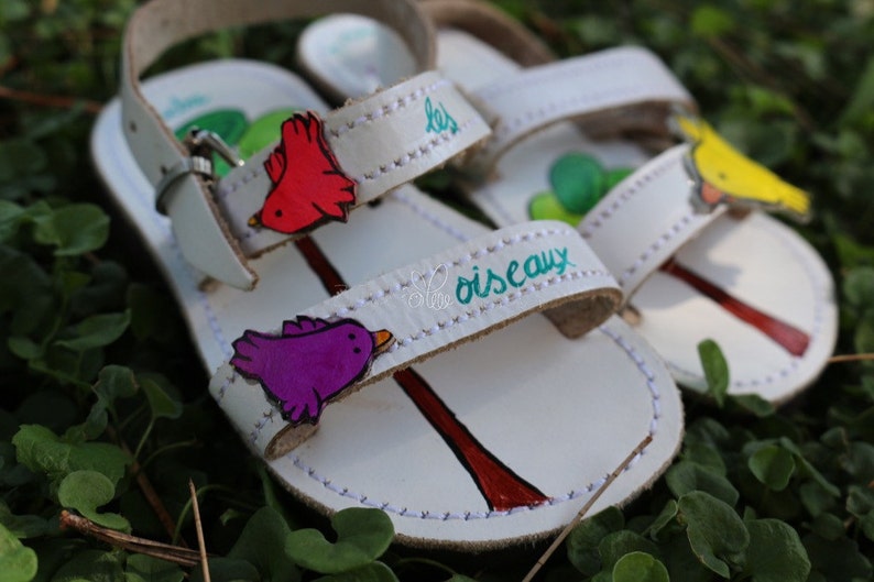 May include: White leather sandals with colourful bird appliques and the text "les oiseaux" on the strap.