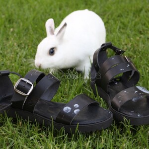May include: A pair of black leather sandals with white dandelion designs on the straps. The sandals are on a green grassy surface with a white rabbit in the background.
