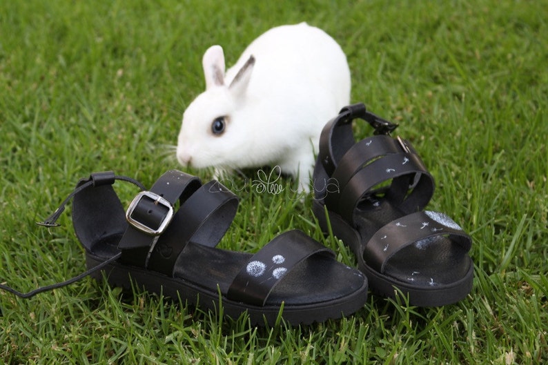 May include: A pair of black leather sandals with white dandelion designs on the straps. The sandals are on a green grassy surface with a white rabbit in the background.