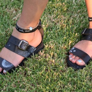May include: Two pairs of black leather sandals with ankle straps. The sandals have white painted designs on them, including a dandelion and a moon. The ankle straps have the text "how dark the night" and "will brush" written on them.