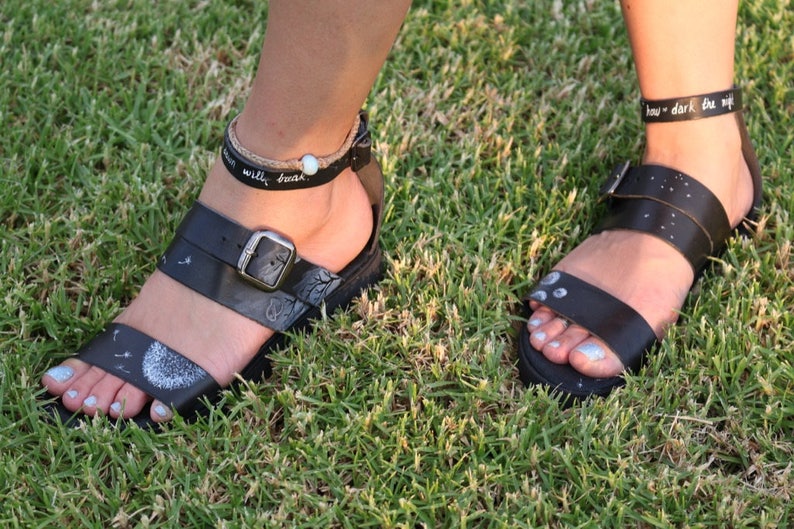 May include: Two pairs of black leather sandals with ankle straps. The sandals have white painted designs on them, including a dandelion and a moon. The ankle straps have the text "how dark the night" and "will brush" written on them.