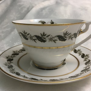 May include: A white teacup and saucer with a black and gold floral design. The cup has a delicate handle and the saucer has a scalloped edge.