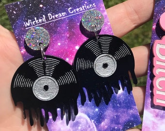 May include: Black vinyl record earrings with silver accents and a dripping design. The earrings have a glittery silver stud and are attached to a purple and blue galaxy-themed card with the text "Wicked Dream Creations".