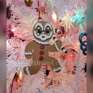 May include: A brown gingerbread man Christmas ornament with white and red accents, holding a candy cane. The ornament has red glitter buttons and is hanging on a white Christmas tree. The text "WickedDreamCreations" is visible.