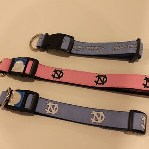 May include: Three dog collars with different colored bands and a black and white logo. The collars are blue, pink, and light blue.