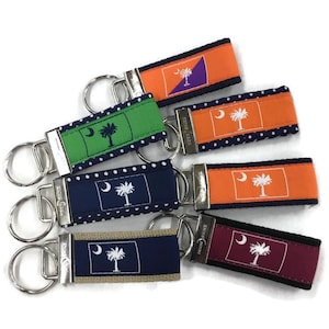 May include: A set of six keychains featuring the South Carolina state flag in various color combinations. The keychains are made of fabric with a white polka dot pattern and a silver metal ring.