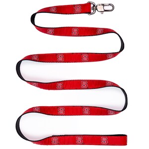 May include: A red fabric lanyard featuring a repeating black and white NC State University logo pattern. The strap has black edges and is finished with a silver-toned metal swivel lobster claw clasp at one end for attaching items securely.