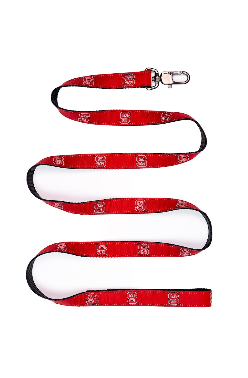 May include: A red fabric lanyard featuring a repeating black and white NC State University logo pattern. The strap has black edges and is finished with a silver-toned metal swivel lobster claw clasp at one end for attaching items securely.