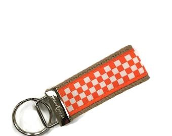 May include: Orange and white checkered key chain with a brown strap and a silver key ring. The key chain has the text "Paddywax" on the silver ring.