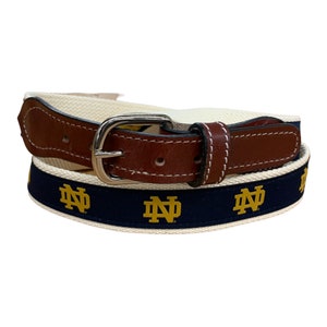 May include: A canvas belt with a brown leather tab and silver buckle. The belt has a navy blue stripe with gold "ND" logos and a cream-colored canvas body.