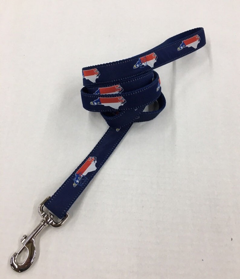 Op de afbeelding: Een blauwe hondenriem met een herhalend patroon van de vlag van de staat North Carolina. De riem heeft een zilveren clip.