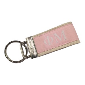 May include: A pink and beige fabric keychain with a silver key ring. The fabric has the letters "PM" embroidered in white.