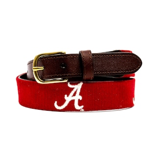 May include: A red and white belt with a brown leather strap and a gold-colored buckle. The belt features the letter "A" in white, likely representing a team or organization. The leather strap is dark brown.