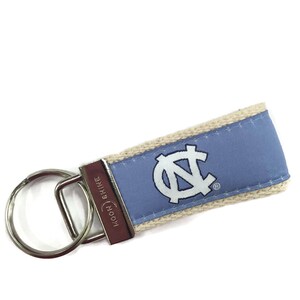 May include: A light blue and white key chain with the University of North Carolina Tar Heels logo. The key chain is made of fabric and has a silver key ring. The key chain is attached to a silver key ring.