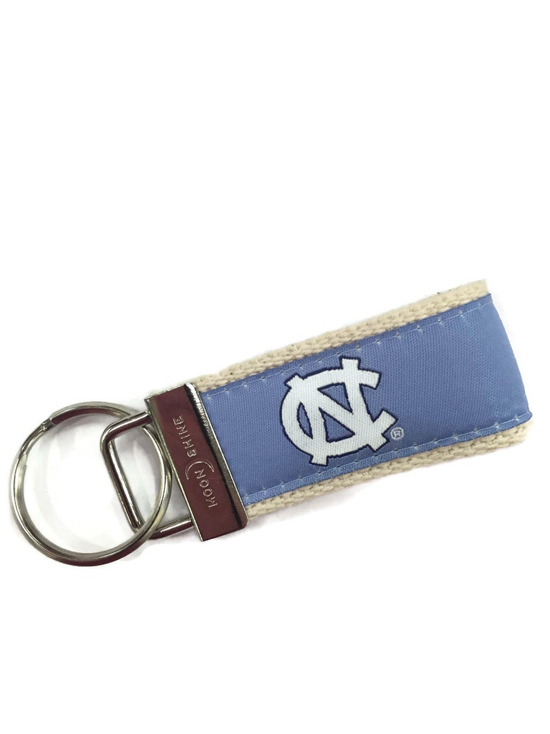 May include: A light blue and white key chain with the University of North Carolina Tar Heels logo. The key chain is made of fabric and has a silver key ring. The key chain is attached to a silver key ring.