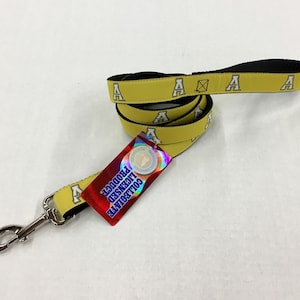 Pode incluir: Trela de cão amarela com uma correia preta e logotipos "A" brancos. A trela tem um clipe prateado e uma etiqueta vermelha que diz "Produto licenciado pela universidade".
