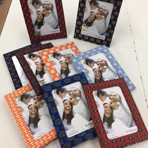 May include: A collection of eight 5x7 inch picture frames with different college team logos. The frames are made of fabric and have a variety of colors and patterns. The frames are all from the same brand, K-Dieter.