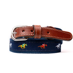 May include: A navy blue belt with a brown leather strap and silver buckle. The belt features embroidered images of jockeys on horses in yellow, red, and white. A classic accessory for equestrian enthusiasts.