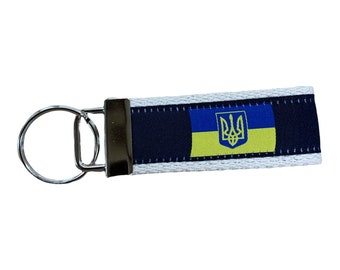 May include: A white, blue and yellow key chain featuring the Ukrainian flag and coat of arms. The key chain is made of fabric and has a silver metal ring.