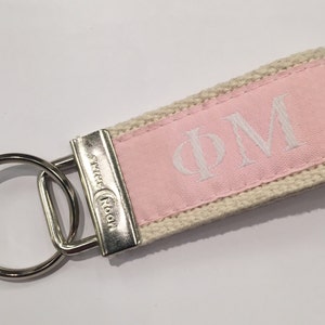 May include: A pink and cream fabric keychain with a silver key ring. The fabric has the initials "PM" embroidered in white.