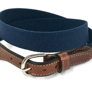May include: A navy blue canvas belt with a brown leather tab and a silver-colored buckle. The leather tab is stitched with white thread. The belt is a classic accessory.