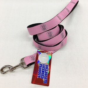 May include: A pink dog leash with a black underside and a repeating logo featuring the letters NC in a collegiate font. The leash includes a silver metal swivel snap hook and a rectangular tag that reads COLLEGIATE LICENSED PRODUCT.