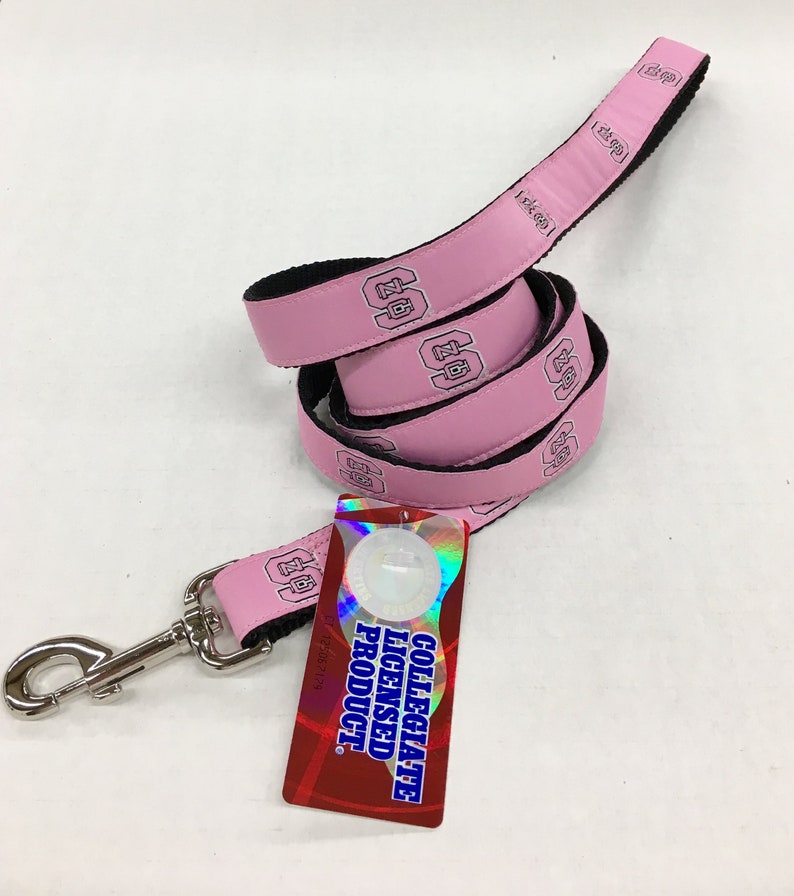 May include: A pink dog leash with a black underside and a repeating logo featuring the letters NC in a collegiate font. The leash includes a silver metal swivel snap hook and a rectangular tag that reads COLLEGIATE LICENSED PRODUCT.