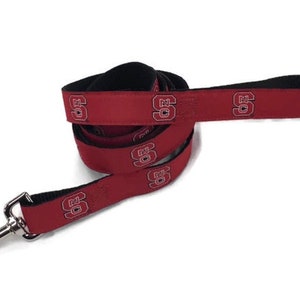 May include: A red dog leash with a black underside and a repeating white and black NC State University logo pattern. The leash features a silver-toned metal swivel snap hook at one end and a looped handle at the other end for secure attachment.