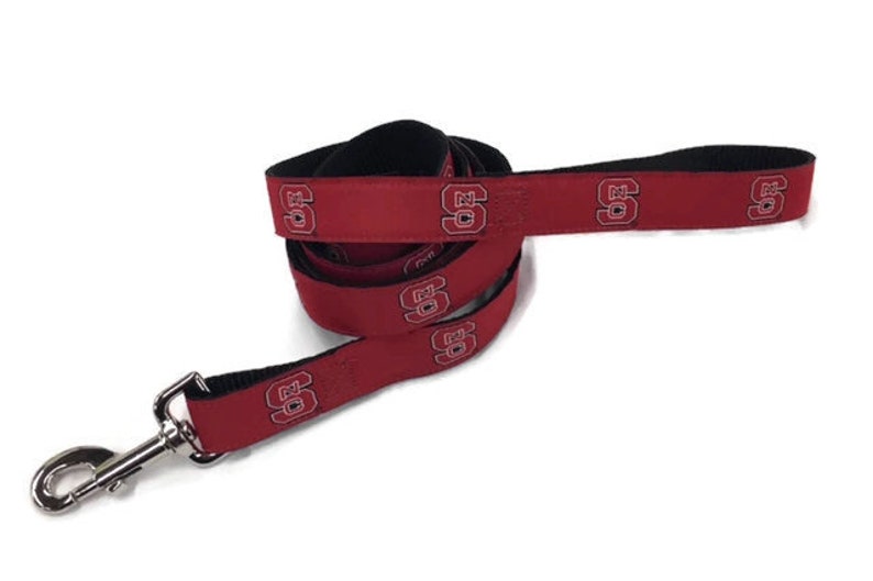 May include: A red dog leash with a black underside and a repeating white and black NC State University logo pattern. The leash features a silver-toned metal swivel snap hook at one end and a looped handle at the other end for secure attachment.