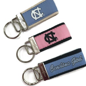 May include: Three keychains with a blue, pink, and light blue fabric strap. Each strap has a white and black UNC logo. The blue strap says "Carolina Girl".