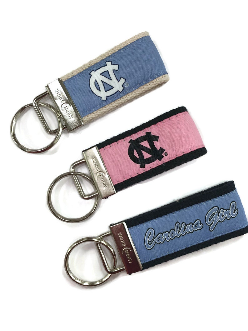 May include: Three keychains with a blue, pink, and light blue fabric strap. Each strap has a white and black UNC logo. The blue strap says "Carolina Girl".
