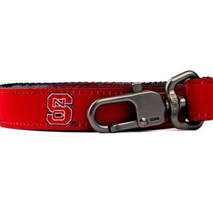May include: A red fabric strap featuring the white and red NC State University logo. The strap includes a dark metallic swivel lobster clasp attached to one end with a rectangular metal loop and reinforced stitching along the edges of the fabric.