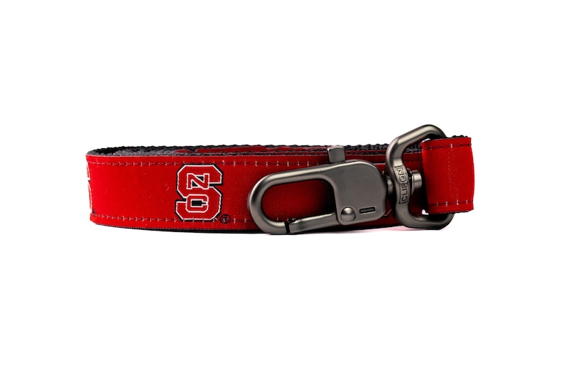 May include: A red fabric strap featuring the white and red NC State University logo. The strap includes a dark metallic swivel lobster clasp attached to one end with a rectangular metal loop and reinforced stitching along the edges of the fabric.