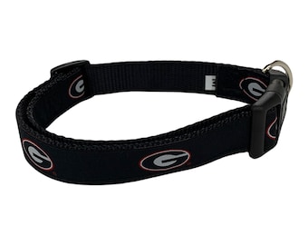 May include: Black dog collar featuring a repeating pattern of the University of Georgia Bulldogs logo in red, white, and gray. The collar has a black plastic buckle and a metal D-ring for leash attachment.