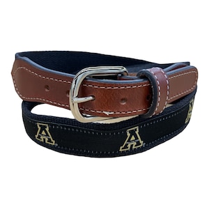 May include: A black and brown leather belt with a silver buckle. The belt has a repeating pattern of gold "A"s on the black canvas strap.