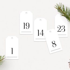 Könnte beinhalten: Weiße Geschenkanhänger mit den schwarzen Zahlen 1, 8, 14, 19 und 23. Die Anhänger sind auf einer weißen Oberfläche mit einem Zweig eines Nadelbaums und einem Tannenzapfen angeordnet. Die Anhänger haben oben ein kleines Loch.