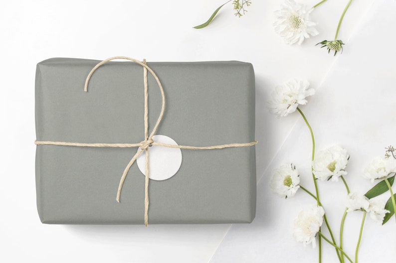 May include: A gray gift box wrapped in brown paper with a white tag tied with twine. The box is surrounded by white flowers with green stems.