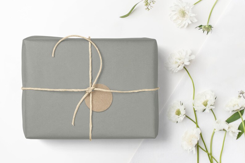 May include: A gray gift box wrapped in brown paper with a brown paper tag and tied with twine. The box is surrounded by white flowers with green stems.