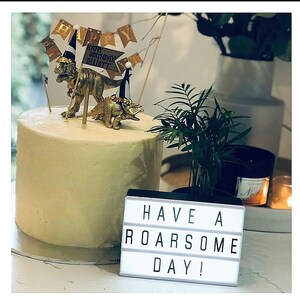 May include: A birthday cake decorated with gold dinosaur figurines and a banner saying "Happy Birthday Elliott." A light-up sign displays "Have a Roarsome Day!" A small plant and candle are also present.