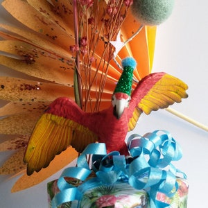 Puede incluir: Una caja de regalo decorada con papel tropical, adornada con una cinta azul brillante. Una figura de loro rojo y amarillo con un gorro de fiesta se encuentra encima de la caja, complementada con flores secas y un elemento de papel naranja en forma de abanico.