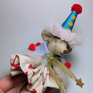 Puede incluir: Una muñeca de ratón de tela hecha a mano vestida con un atuendo festivo. El ratón lleva un gorro de fiesta azul con lunares dorados y un pompón rojo. Su atuendo incluye una capa azul claro, una falda crema con forma de corazón rojo y detalles dorados en forma de estrella.