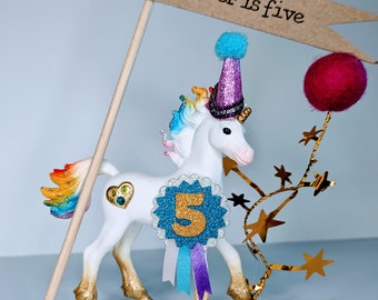 Schleich rainbow unicorn cake topper wearing a party hat and badge, includes a mini flag with wording of your choice. Add extra accessories.