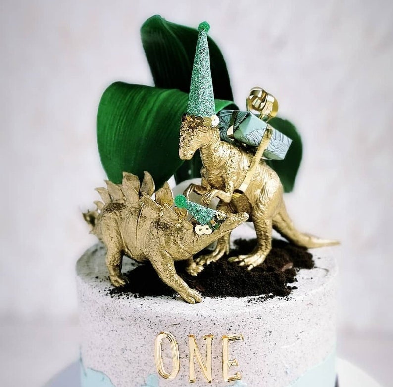 May include: A gold dinosaur cake topper with two dinosaurs wearing party hats and carrying gifts. The cake is decorated with the word "ONE" in gold lettering and a large green leaf.