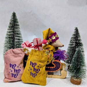 Puede incluir: Una escena festiva navideña con un juguete de peluche con un sombrero adornado con estrellas, sentado en un trineo de madera. Dos pequeños árboles de Navidad flanquean la escena. También hay varias bolsas de regalo en rosa y dorado, impresas con "ho ho ho".