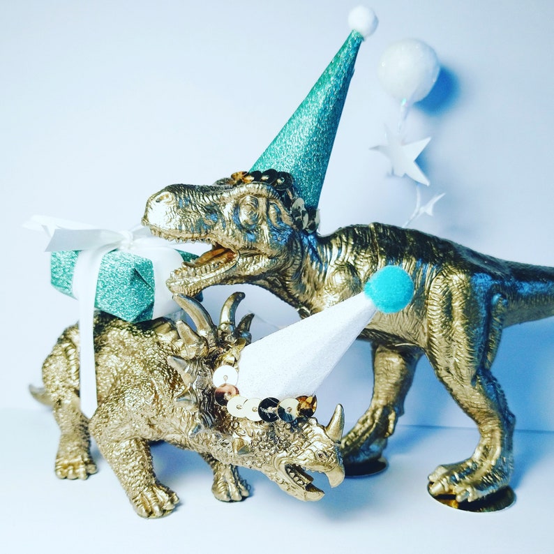 May include: Two gold dinosaur figurines, a Tyrannosaurus Rex and a Triceratops, wear party hats and sequins. The T-Rex holds a teal gift box tied with a white ribbon. Decorative stars and a balloon are in the background.