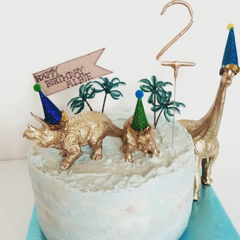 May include: A birthday cake featuring gold dinosaur decorations and a number 2 candle. The cake is topped with a banner reading "HAPPY BIRTHDAY ALBIE". The dinosaurs wear party hats, and the cake has blue and white frosting.