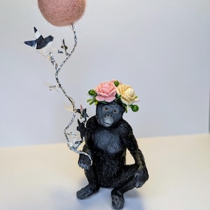 Op de afbeelding: Een zwart beeldje van een chimpansee met een bloemenkrans en een roze ballon met zilveren sterren.