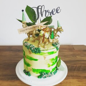 May include: A birthday cake with green and yellow frosting, decorated with gold dinosaur figurines and a "Three" cake topper. The cake has a "Happy Birthday Parker" banner and is on a white plate.