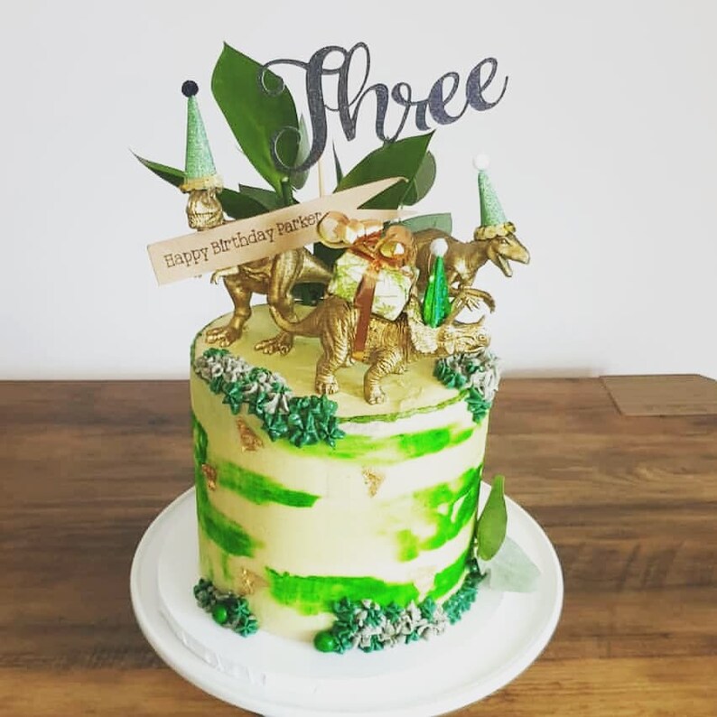 May include: A birthday cake with green and yellow frosting, decorated with gold dinosaur figurines and a "Three" cake topper. The cake has a "Happy Birthday Parker" banner and is on a white plate.