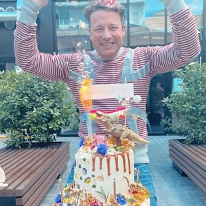 May include: A birthday cake decorated with a dinosaur theme, featuring brown frosting drips, colourful flowers, and a toy dinosaur. The cake is topped with candles and a sign that reads "The Naked Chef". A person is wearing a red and white striped shirt and a party hat.